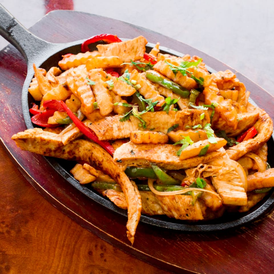 Sizzling chicken fajitas with peppers, onions, and fries in a cast iron skillet on a wooden board.