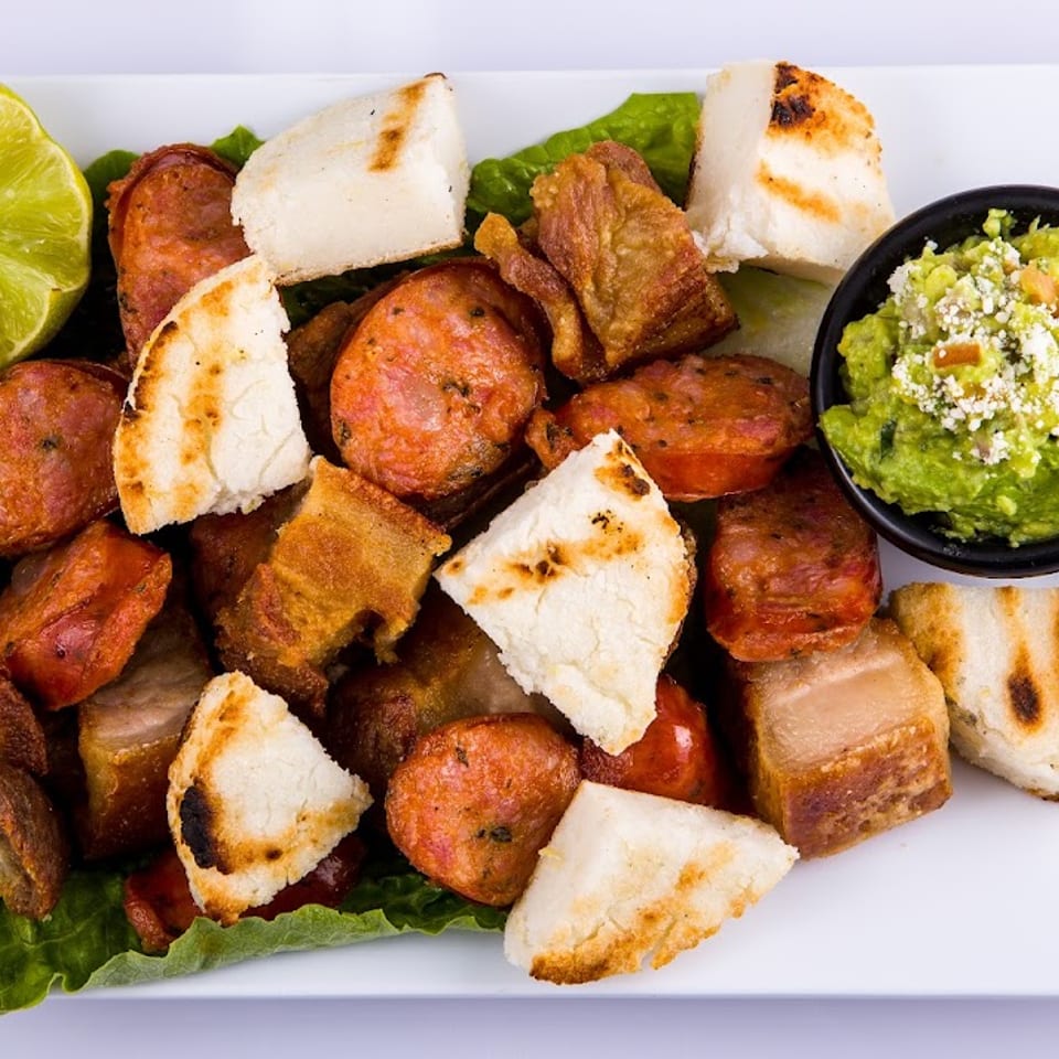 A white plate with fried pork, chorizo, grilled arepas, lime, and guacamole on lettuce.